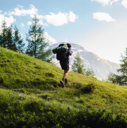 A man in black boots with a black bag walking on green grass wearing waterproof socks is ready for a hike.