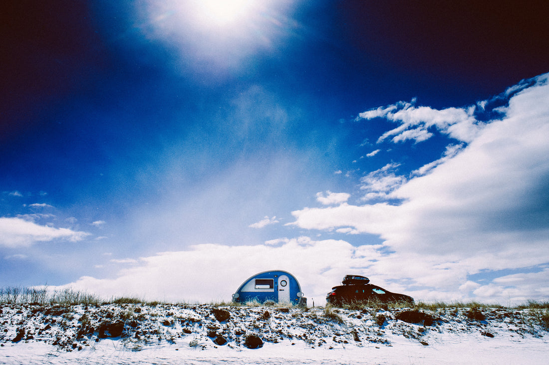 A blue and white teardrop camper is towed by a sedan along a road lined with snow, under a bright blue sky near Boulder, Utah