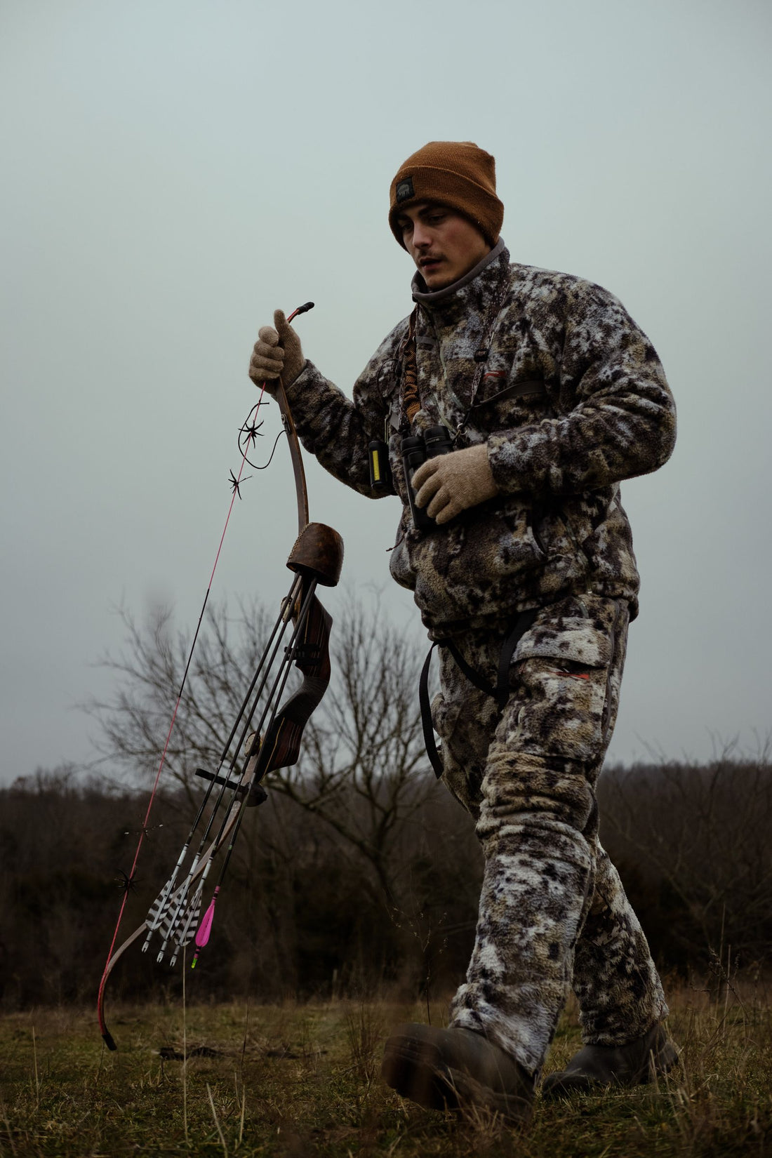 Bowhunter in full camouflage carrying a bow and arrows across an open field during deer season