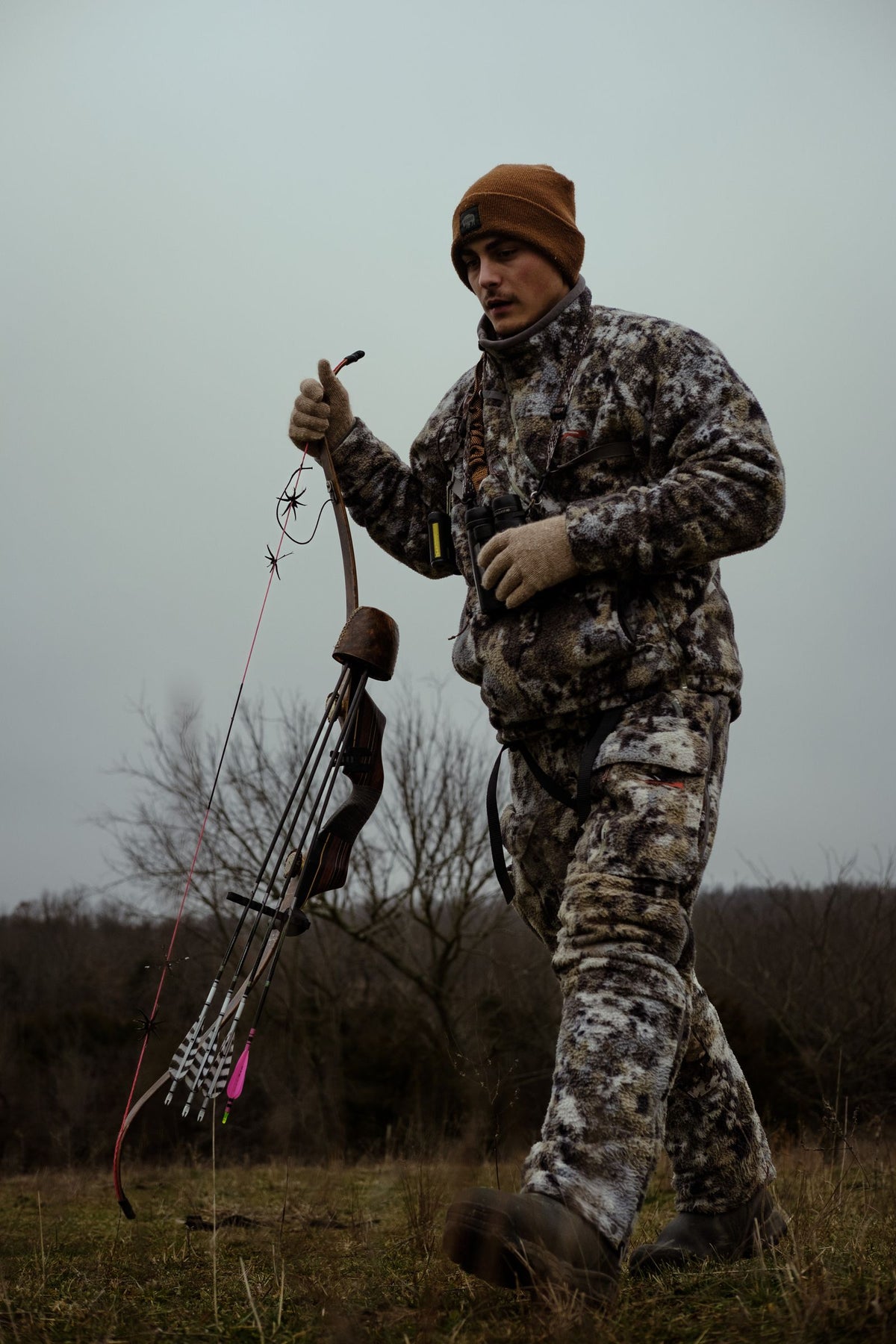 Bowhunter in full camouflage carrying a bow and arrows across an open field during deer season