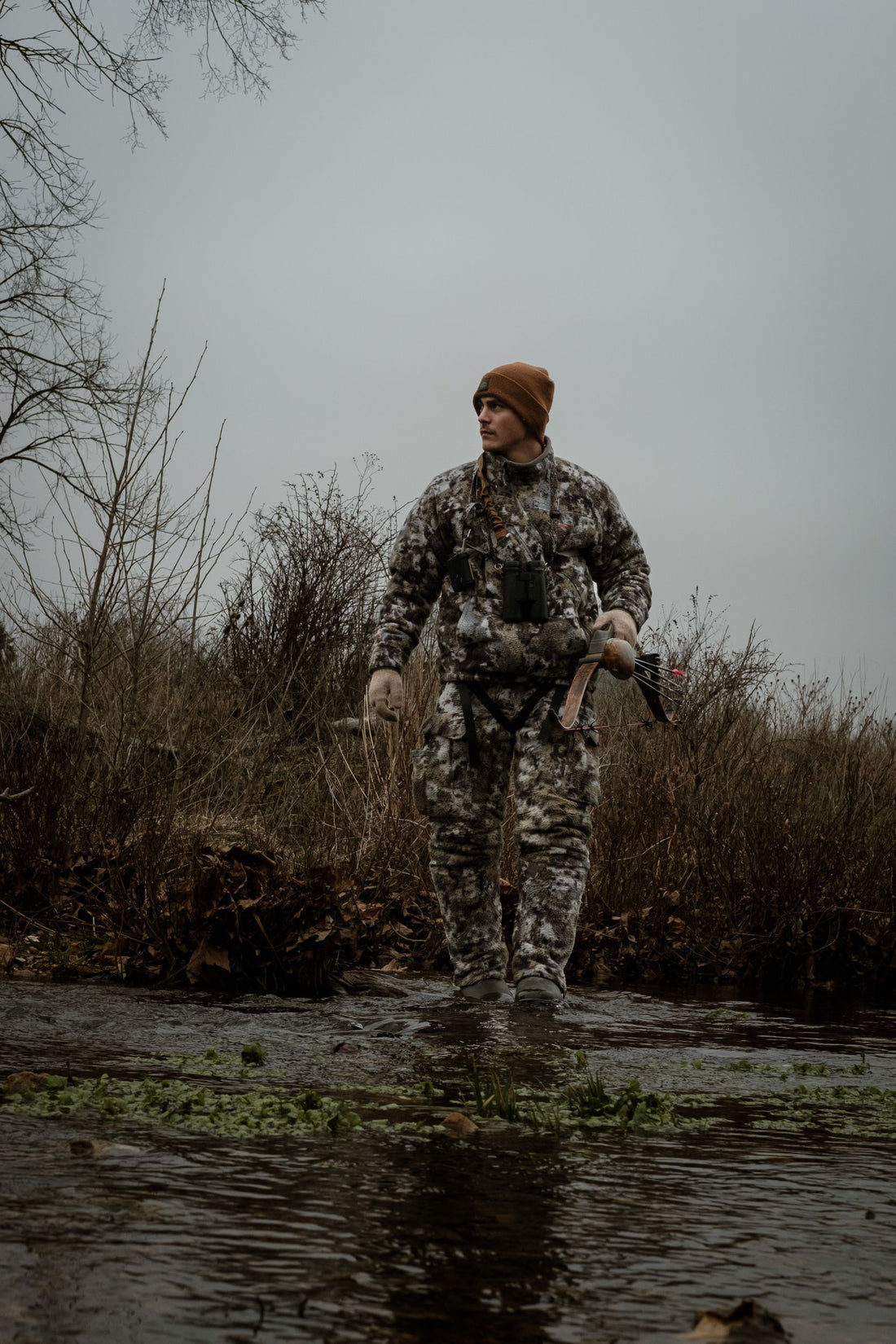 Deer hunter in full camouflage carrying a bow while wading across a creek in autumn woods