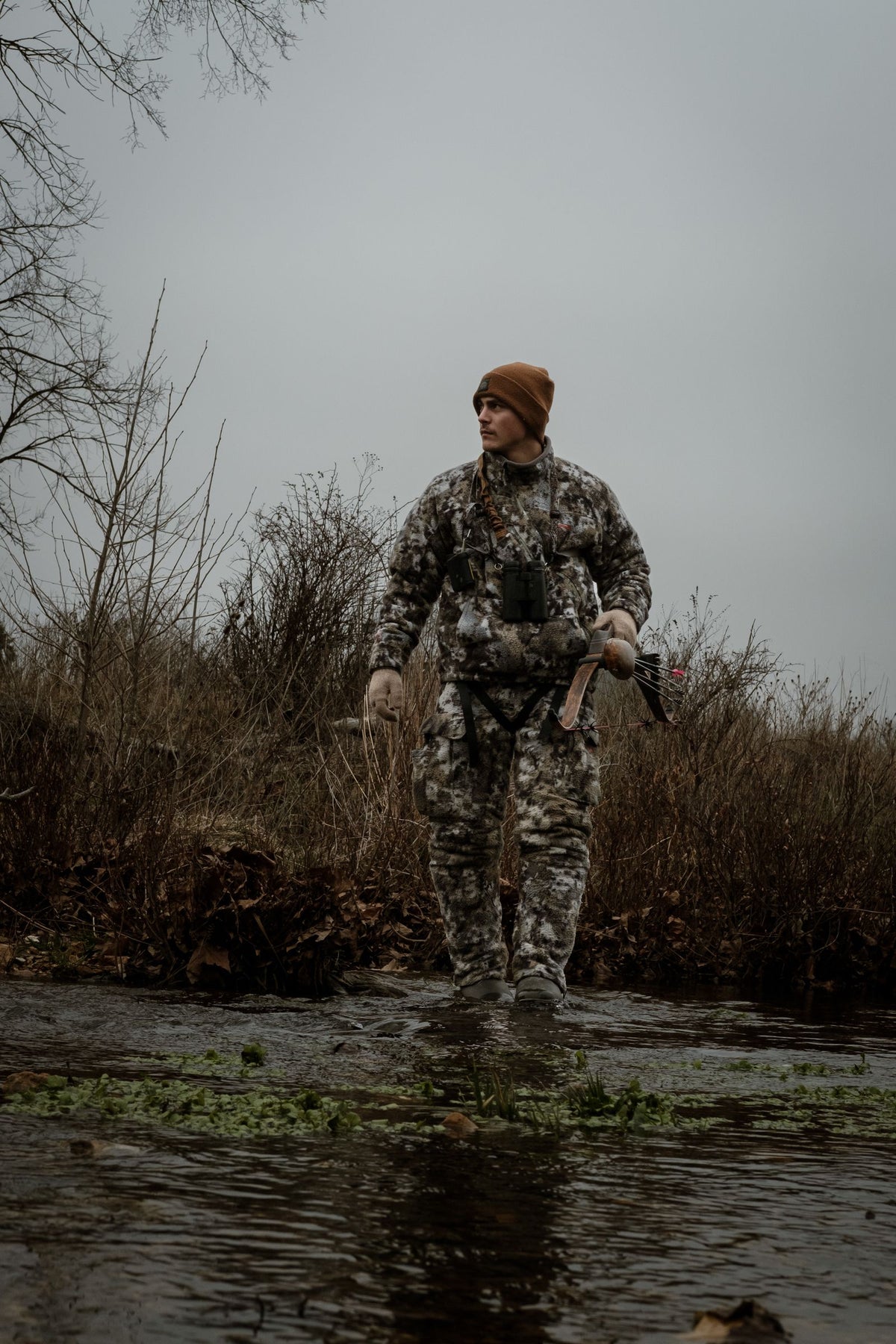 Deer hunter in full camouflage carrying a bow while wading across a creek in autumn woods