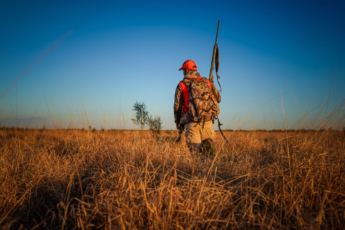 Deer hunter in camouflage and a hunter orange cap walking away across an open field with a rifle